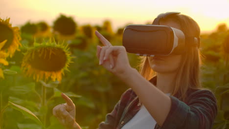 The-woman-with-long-hair-in-plaid-shirt-and-jeans-is-working-in-VR-glasses.-She-is-engaged-in-the-working-process.-It-is-a-great-sunny-evening-in-the-sunflower-field-at-sunset.